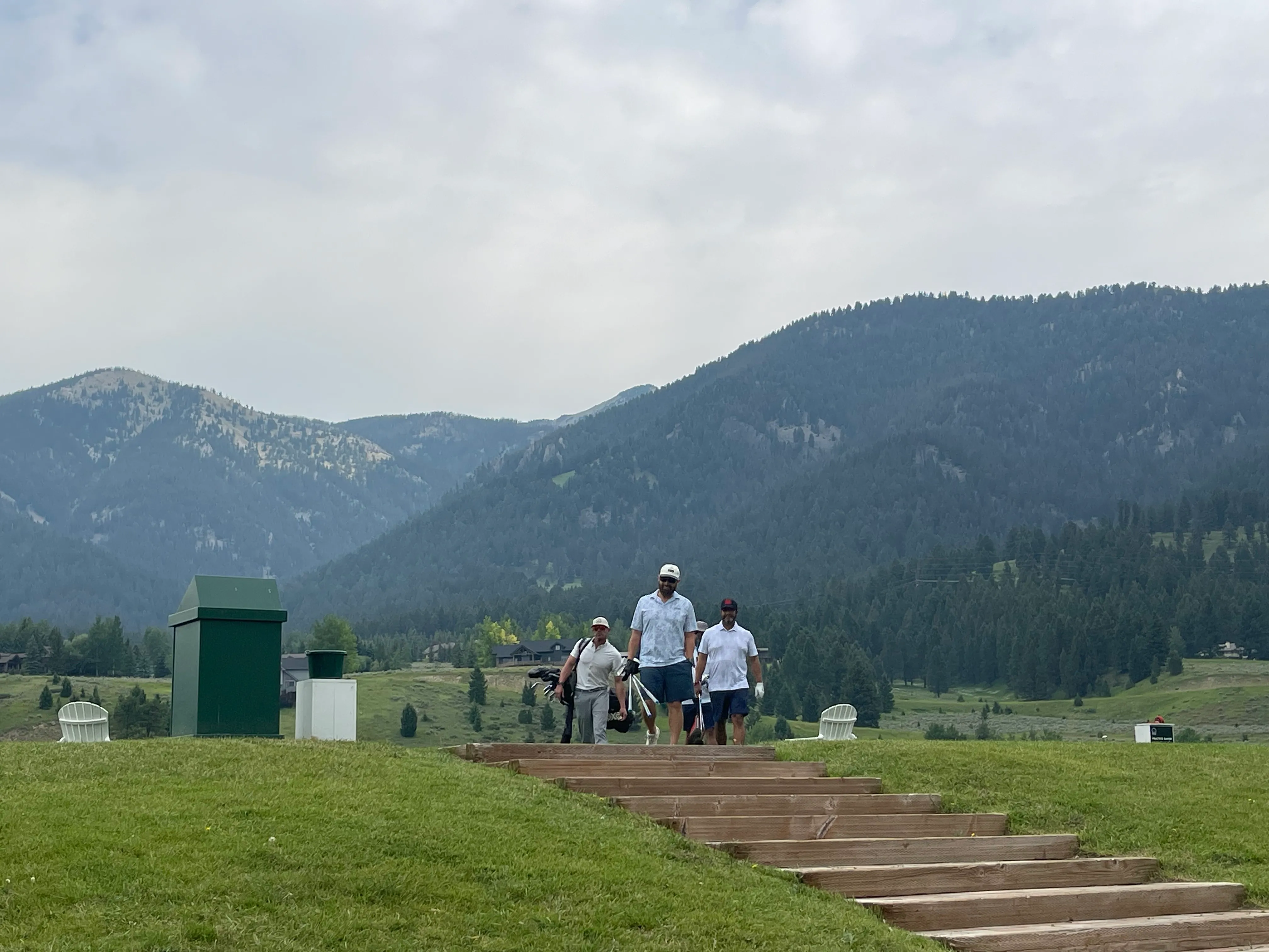 Three golfers carrying their bags down wooden steps with mountains behind them.