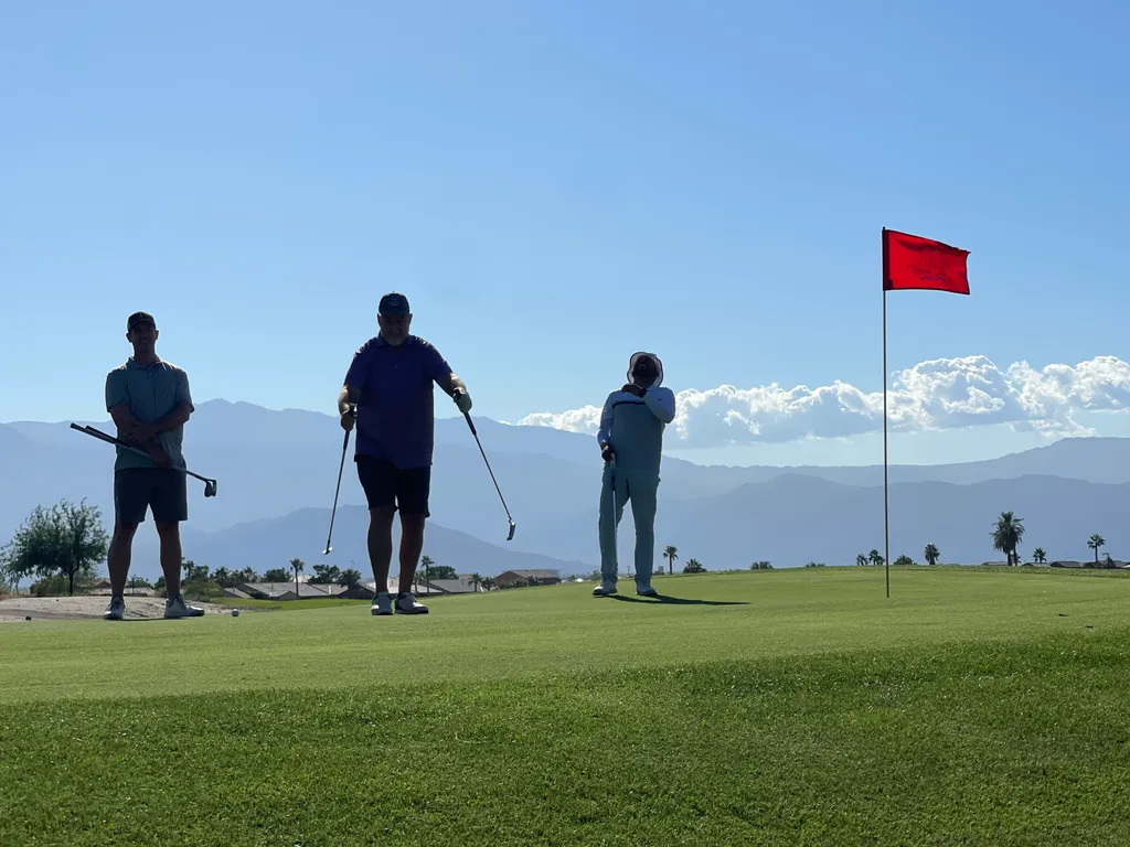 Three golfers on a putting green beside a red flag, with mountains behind them.